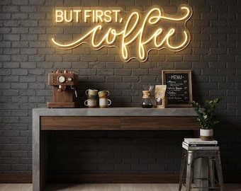 May include: A glowing neon sign on a dark brick wall displays the text "BUT FIRST, coffee" in a warm yellow script. Below, a wooden coffee machine, several small ceramic mugs, a glass pour-over carafe, a menu board, and a potted green plant.