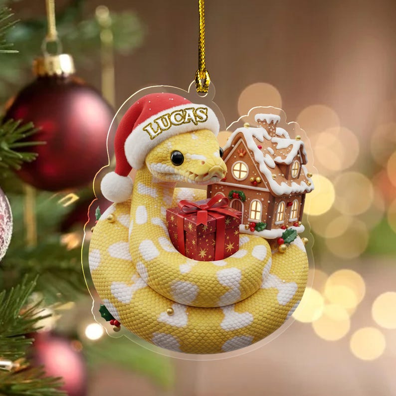May include: A Christmas tree ornament depicting a yellow snake wearing a Santa hat with the name "LUCAS". The snake is coiled around a red gift box and a gingerbread house. The ornament is suspended from a Christmas tree.