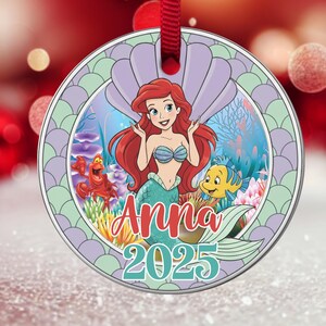 May include: Round Christmas ornament featuring Ariel from The Little Mermaid. The ornament has a red ribbon for hanging. Ariel is depicted with red hair, a blue seashell top, and a green tail. The ornament has the name "Anna" and the year "2025".