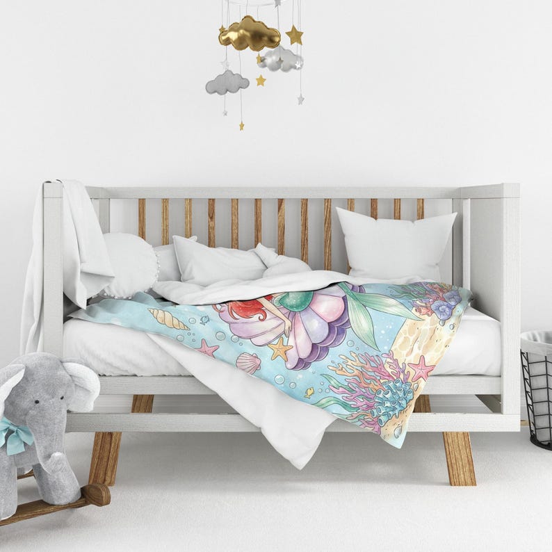 K&ouml;nnte beinhalten: Eine Babydecke mit einem Meerjungfrau-Design. Die Decke hat einen hellblauen Hintergrund mit Illustrationen von Muscheln, Seesternen und Korallen. Die Meerjungfrau hat rotes Haar und einen gr&uuml;nen Schwanz. Die Decke liegt auf einem wei&szlig;en Kinderbett.