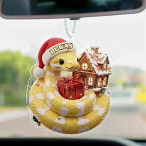 May include: A festive Christmas tree ornament featuring a yellow and white snake wearing a Santa hat with the name "LUCAS". The snake is coiled around a red gift and a gingerbread house, suspended by a silver chain.