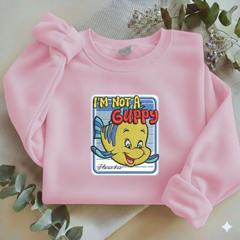 May include: A light pink crewneck sweatshirt featuring a central embroidered patch of a yellow and blue fish character. The text above the fish reads I'M NOT A GUPPY in bold red and yellow letters, with the name Flounder written in cursive below.