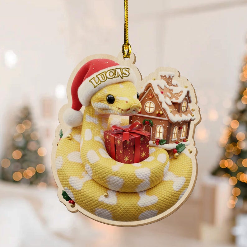 May include: Wooden Christmas decoration featuring a yellow snake wearing a Santa hat with the name "LUCAS". The snake is coiled around a red gift box and a gingerbread house. The decoration has a gold string for hanging.