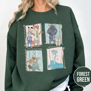 K&ouml;nnte beinhalten: Forstgr&uuml;nes Sweatshirt mit vier gerahmten Bildern von Zootopia-Charakteren. Die Charaktere sind Judy Hopps, Nick Wilde, Chief Bogo, Flash und andere. Das Sweatshirt hat eine einfarbige forstgr&uuml;ne Farbe.
