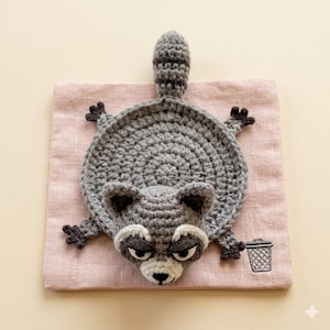 May include: A handmade crochet raccoon, primarily gray with white and black accents, lies on a pink square. The raccoon has a circular body, a striped tail, and small black paws. A small embroidered trash can is in the lower right corner.