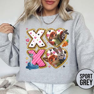 May include: Sport grey sweatshirt featuring a colorful graphic design. The design includes the letters "XO" with heart and character illustrations. The sweatshirt has a crew neck and long sleeves. The text "SPORT GREY" is printed on a circular label.