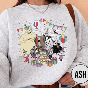 May include: A light gray sweatshirt featuring a colorful graphic of characters from The Nightmare Before Christmas celebrating a birthday. The design includes a cake, balloons, and the number 4. The characters are surrounded by colorful party decorations.
