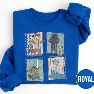 K&ouml;nnte beinhalten: K&ouml;nigsblaues Sweatshirt mit vier illustrierten Panels von Zeichentrickfiguren. Die Panels zeigen Judy Hopps und Nick Wilde, Chief Bogo und Flash, einen Biber und eine Schlange. Das Wort "ROYAL" ist auf einem blauen Kreis aufgedruckt.