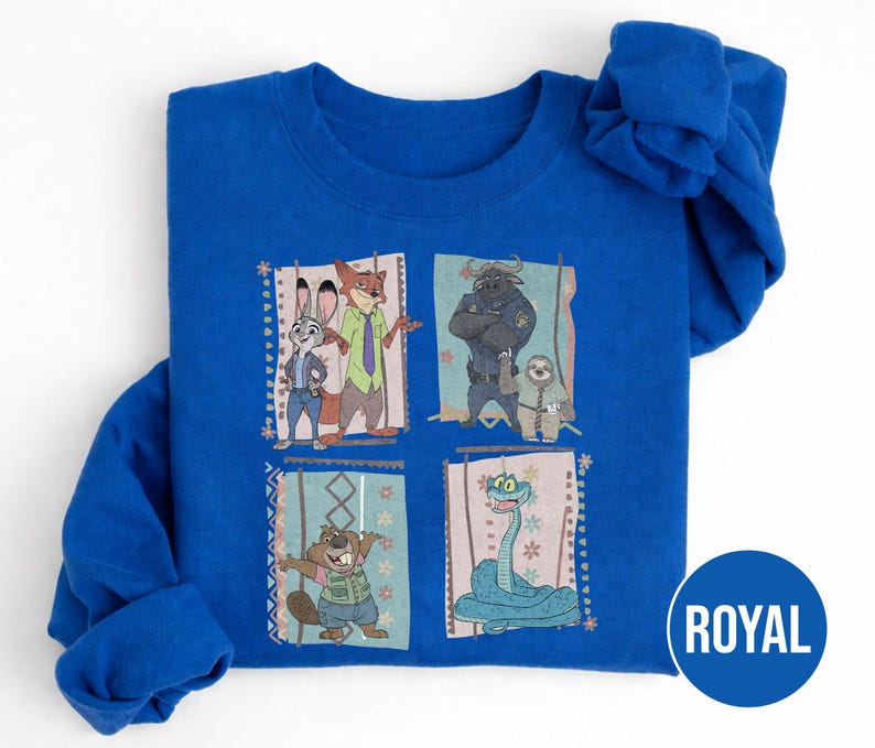 K&ouml;nnte beinhalten: K&ouml;nigsblaues Sweatshirt mit vier illustrierten Panels von Zeichentrickfiguren. Die Panels zeigen Judy Hopps und Nick Wilde, Chief Bogo und Flash, einen Biber und eine Schlange. Das Wort "ROYAL" ist auf einem blauen Kreis aufgedruckt.