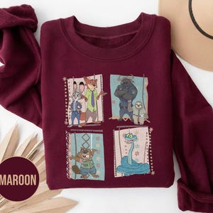 K&ouml;nnte beinhalten: Ein weinrotes Sweatshirt mit einem farbenfrohen Grafikdesign von Zeichentrickfiguren. Die Grafik ist in vier Felder unterteilt, die jeweils eine andere Figur zeigen. Das Wort "MAROON" ist auf einem kreisf&ouml;rmigen Etikett aufgedruckt.