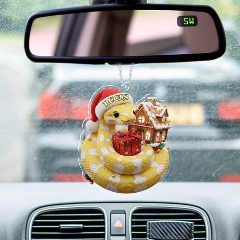 May include: A festive car air freshener featuring a yellow and white snake wearing a Santa hat with the name "LUCAS". It holds a red gift and is next to a gingerbread house. The ornament hangs from a car's rearview mirror.