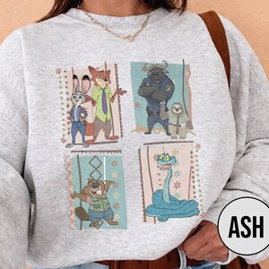 K&ouml;nnte beinhalten: Ein hellgraues Sweatshirt mit einem farbenfrohen Grafikdesign von Zeichentrickfiguren. Das Design umfasst vier rechteckige Tafeln mit Bildern eines Kaninchens, Fuchses, Bullen, Faultiers, Bibers und einer Schlange. Das Wort "ASH" befindet sich in einem Kreis unten rechts.