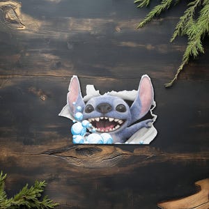 Puede incluir: Un personaje de dibujos animados azul, Stitch, irrumpe a trav&eacute;s de una superficie blanca rasgada, rodeado de burbujas azules. Stitch tiene orejas grandes, una amplia sonrisa y est&aacute; sobre un fondo de madera oscura con ramitas verdes.