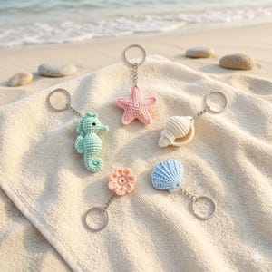 May include: A collection of crocheted keychains in the shape of a seahorse, starfish, seashell, flower, and clam shell. The keychains are in pastel colors: light blue, pink, white, and peach. The keychains are on a white towel, with a beach background.