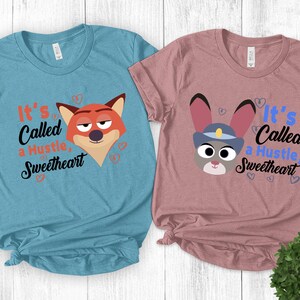 K&ouml;nnte beinhalten: Zwei T-Shirts, eines hellblau und eines mauve, jeweils mit einem Cartoon-Tiergesicht und dem Text "It's Called a Hustle, Sweetheart". Das hellblaue Shirt hat einen Fuchs, und das mauvefarbene Shirt hat ein Kaninchen.