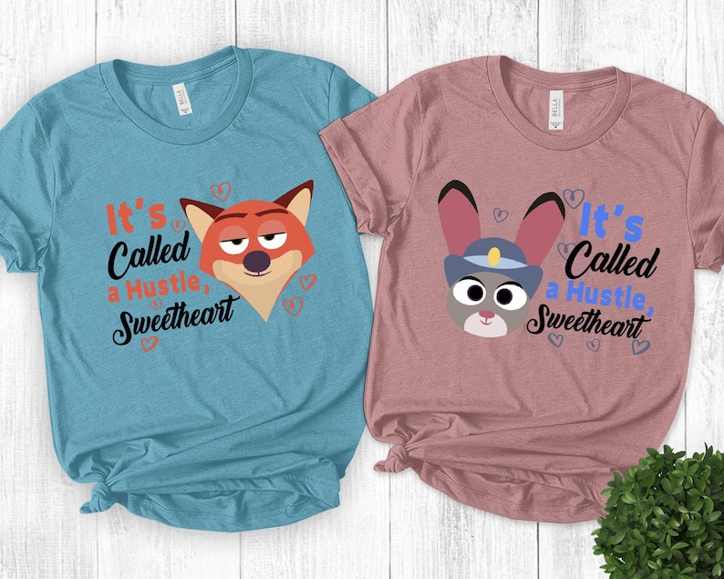 K&ouml;nnte beinhalten: Zwei T-Shirts, eines hellblau und eines mauve, jeweils mit einem Cartoon-Tiergesicht und dem Text "It's Called a Hustle, Sweetheart". Das hellblaue Shirt hat einen Fuchs, und das mauvefarbene Shirt hat ein Kaninchen.