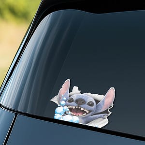Puede incluir: Una calcoman&iacute;a azul y blanca del personaje Stitch de Lilo & Stitch, rompiendo una superficie rasgada. El personaje tiene orejas grandes, una amplia sonrisa y burbujas azules.