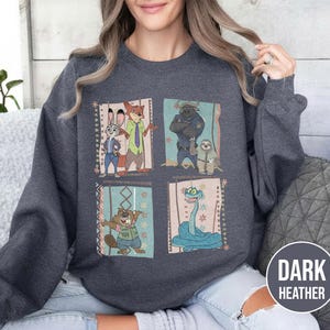 K&ouml;nnte beinhalten: Dunkelgraues Sweatshirt mit einem farbenfrohen Grafikdesign von Zeichentrickfiguren. Das Design umfasst vier rechteckige Tafeln mit verschiedenen Charakteren. Das Sweatshirt hat einen Rundhalsausschnitt und lange &Auml;rmel.