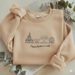 May include: A beige crewneck sweatshirt featuring a black line art graphic of a castle, a mouse ear icon, a large tree, and a geodesic dome. Below the illustrations, the text reads "Happiest place on earth" in a black cursive script font.