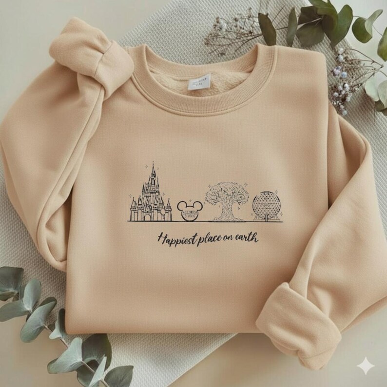 May include: A beige crewneck sweatshirt featuring a black line art graphic of a castle, a mouse ear icon, a large tree, and a geodesic dome. Below the illustrations, the text reads "Happiest place on earth" in a black cursive script font.