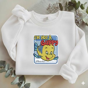 May include: A white crewneck sweatshirt featuring a central embroidered graphic of a yellow fish with blue fins. The text reads I'M NOT A GUPPY in yellow and red, with the name Flounder written in cursive script below the fish inside a blue border.