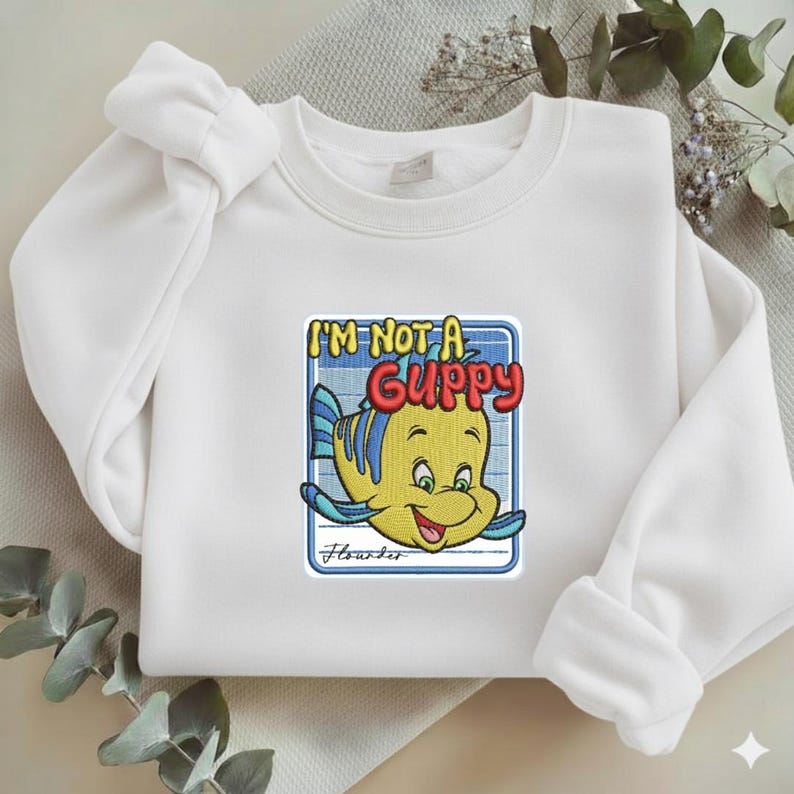 May include: A white crewneck sweatshirt featuring a central embroidered graphic of a yellow fish with blue fins. The text reads I'M NOT A GUPPY in yellow and red, with the name Flounder written in cursive script below the fish inside a blue border.