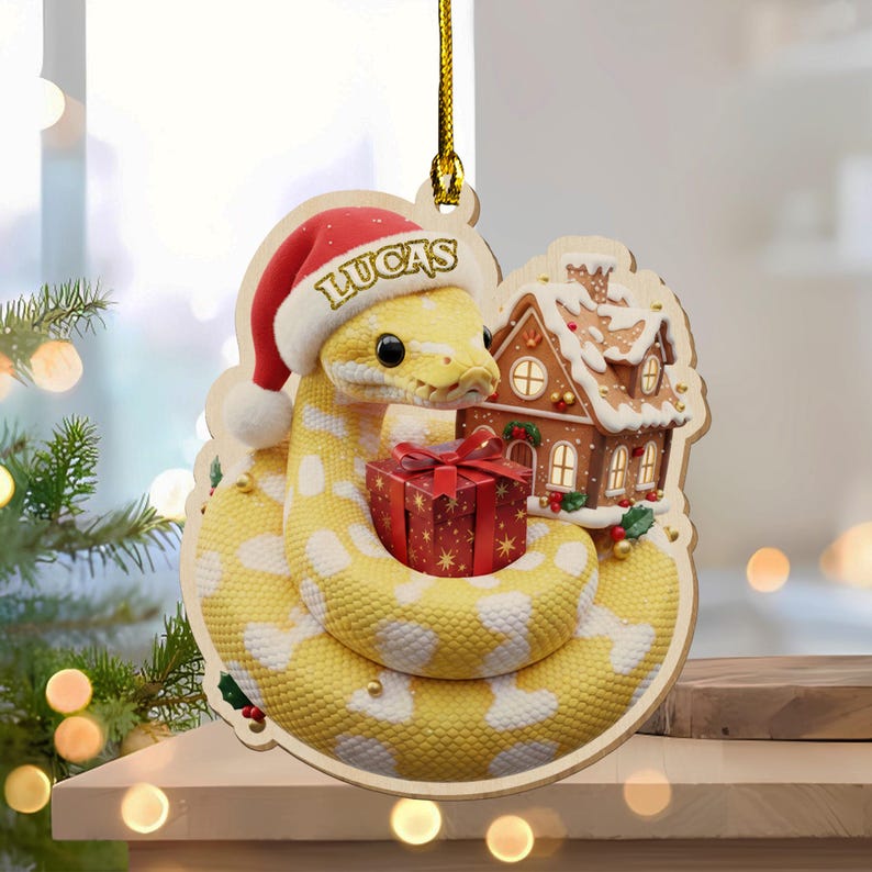 May include: A wooden Christmas ornament depicting a yellow snake with white spots wearing a Santa hat with the name "LUCAS". The snake is coiled around a red gift box and a gingerbread house. The ornament hangs from a gold string.