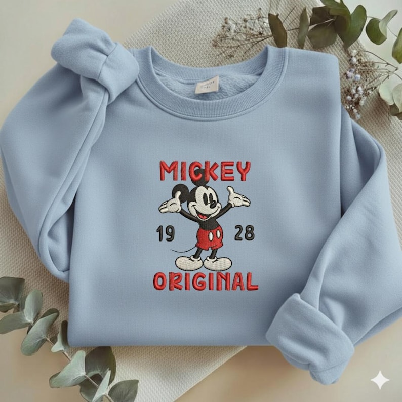 May include: A light blue crewneck sweatshirt featuring an embroidered graphic of Mickey Mouse in the center. The text "MICKEY" appears above the character, "19 28" is on the sides, and "ORIGINAL" is below, all in red stitched lettering.