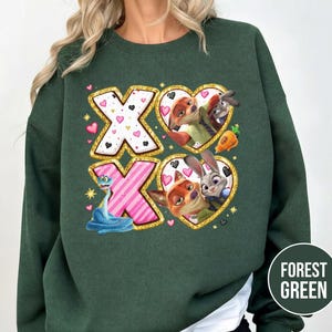 May include: Forest green sweatshirt featuring a colorful graphic with the text "XO". The graphic includes images of cartoon characters, hearts, and a snake. The design is outlined in gold glitter.