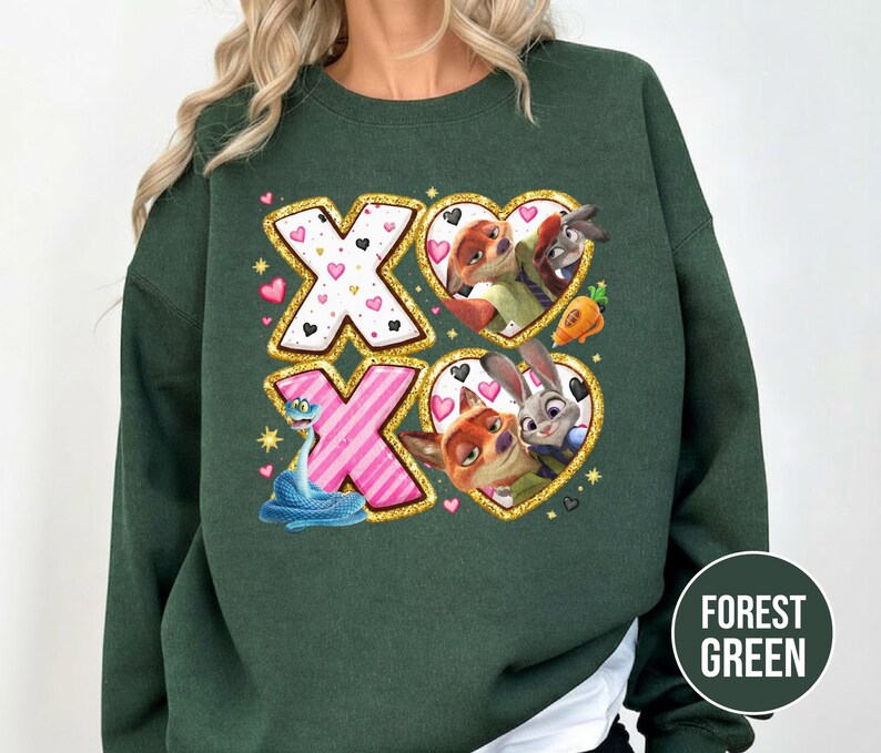 May include: Forest green sweatshirt featuring a colorful graphic with the text "XO". The graphic includes images of cartoon characters, hearts, and a snake. The design is outlined in gold glitter.