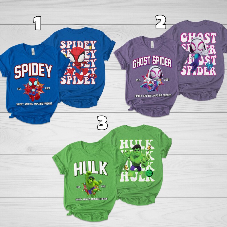 Puede incluir: Tres camisetas de manga corta con dise&ntilde;os de superh&eacute;roes. Las camisetas son azules, moradas y verdes. Las camisetas azules dicen "Spidey" con un gr&aacute;fico de Spider-Man. Las camisetas moradas dicen "Ghost Spider". Las camisetas verdes dicen "Hulk".