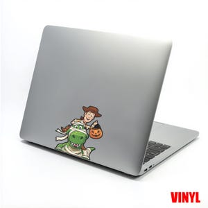May include: A silver laptop with a colorful vinyl sticker featuring Woody from Toy Story riding a mummy-wrapped Rex, holding a jack-o'-lantern. The word "VINYL" is printed in red at the bottom right corner.
