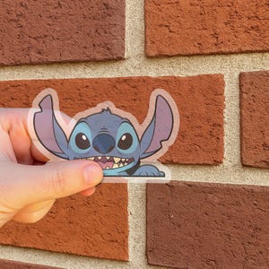 May include: A transparent sticker featuring the character Stitch from the Disney movie Lilo & Stitch. The sticker shows Stitch's face with a wide grin, large eyes, and prominent ears. The sticker is held against a red brick wall.
