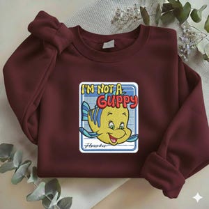 May include: A maroon long-sleeve sweatshirt featuring a central embroidered patch of a yellow and blue fish character. The text reads I'M NOT A GUPPY in yellow and red letters, with the name Flounder written in cursive script below the fish.