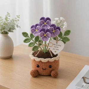 May include: A crocheted flower pot with a smiling face, holding a bouquet of purple pansies, white flowers, and green leaves. A heart-shaped tag tied with twine reads "Keep growing and be your best."