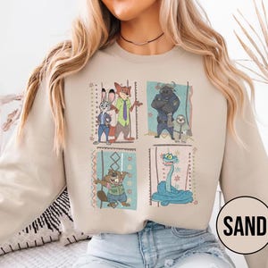 K&ouml;nnte beinhalten: Sandfarbenes Sweatshirt mit vier gerahmten Cartoon-Bildern von Charakteren. Die Bilder zeigen ein Kaninchen, einen Fuchs, einen B&uuml;ffel, ein Faultier, einen Biber und eine Schlange. Das Wort "SAND" ist in einem wei&szlig;en Kreis unten rechts aufgedruckt.