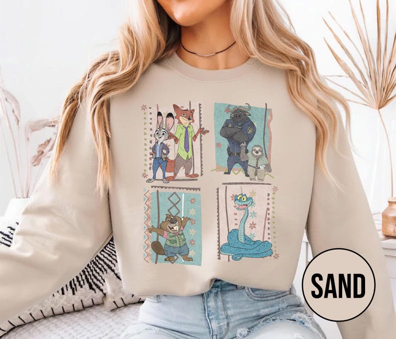 K&ouml;nnte beinhalten: Sandfarbenes Sweatshirt mit vier gerahmten Cartoon-Bildern von Charakteren. Die Bilder zeigen ein Kaninchen, einen Fuchs, einen B&uuml;ffel, ein Faultier, einen Biber und eine Schlange. Das Wort "SAND" ist in einem wei&szlig;en Kreis unten rechts aufgedruckt.