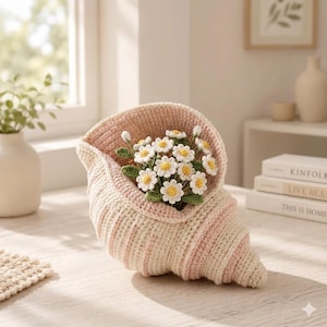 May include: A handcrafted crochet seashell-shaped vase in cream and pink stripes, holding a bouquet of white and yellow daisy-like flowers with green leaves. The vase is on a light-colored surface.