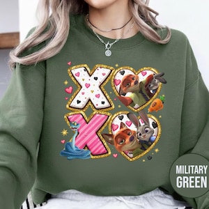 May include: Military green sweatshirt featuring a graphic design with the letters "XO" and heart-shaped frames with cartoon characters. The "X" letters are white with pink hearts, and the "O" letters are pink with black hearts. The sweatshirt has a round neck.