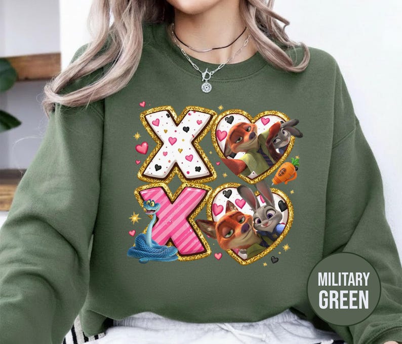 May include: Military green sweatshirt featuring a graphic design with the letters "XO" and heart-shaped frames with cartoon characters. The "X" letters are white with pink hearts, and the "O" letters are pink with black hearts. The sweatshirt has a round neck.