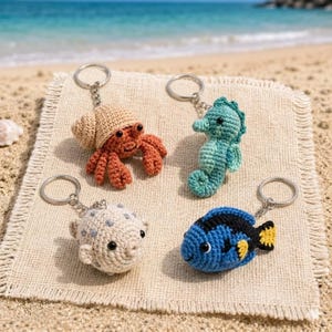 May include: Four crochet keychains featuring sea creatures: a hermit crab, seahorse, pufferfish, and a blue fish with yellow accents. Each keychain is attached to a silver ring. The keychains are displayed on a beige, textured surface with a beach background.