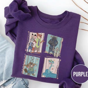 K&ouml;nnte beinhalten: Ein lila Sweatshirt mit einer farbenfrohen Grafik von Charakteren aus dem Animationsfilm Zoomania. Das Design umfasst Judy Hopps, Nick Wilde, Chief Bogo, Flash und andere. Das Wort "PURPLE" befindet sich in einem lila Kreis.