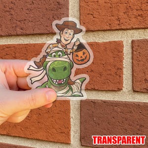May include: Transparent sticker featuring Woody from Toy Story riding a mummy-wrapped Rex, holding a jack-o'-lantern. The sticker has a Halloween theme. The word "TRANSPARENT" is printed at the bottom.