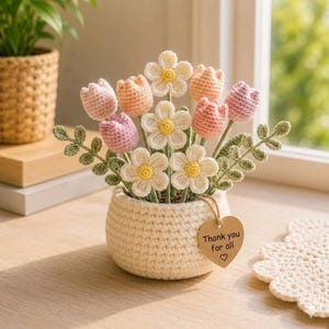 May include: A crocheted bouquet of flowers in a cream-colored pot. The flowers include pink, peach, and white daisies and tulips with green leaves. A heart-shaped tag reads "Thank you for all".