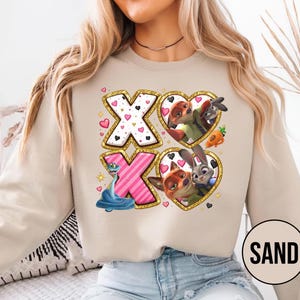 May include: Sand-colored sweatshirt featuring a graphic design with the letters "X" and "O" in gold glitter frames. The "X" letters have heart and polka dot patterns. The "O" letters contain cartoon characters in heart-shaped frames. The word "SAND" is in a circle.