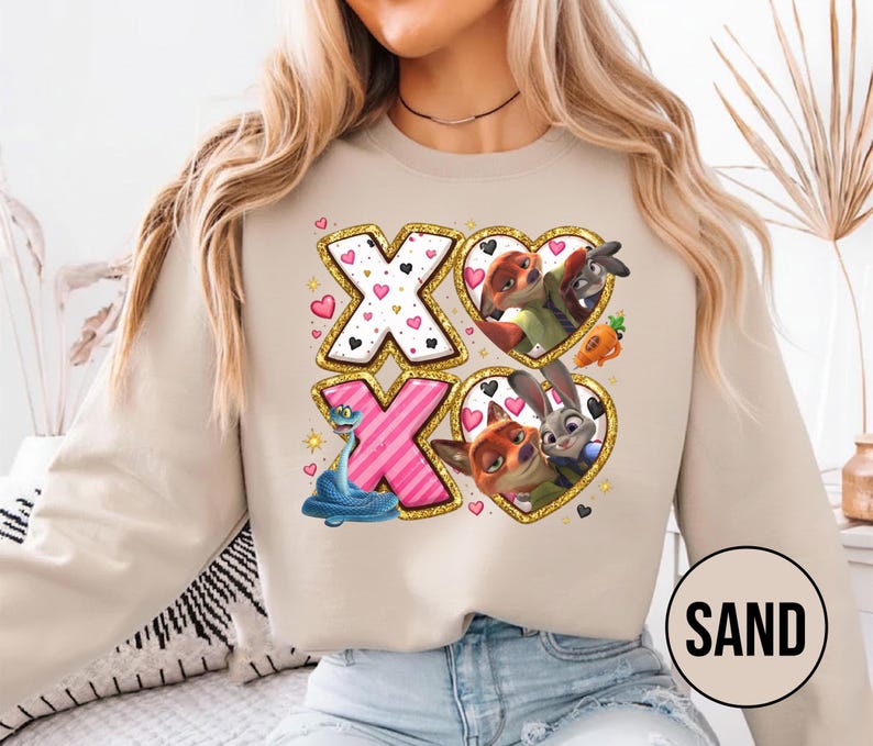 May include: Sand-colored sweatshirt featuring a graphic design with the letters "X" and "O" in gold glitter frames. The "X" letters have heart and polka dot patterns. The "O" letters contain cartoon characters in heart-shaped frames. The word "SAND" is in a circle.