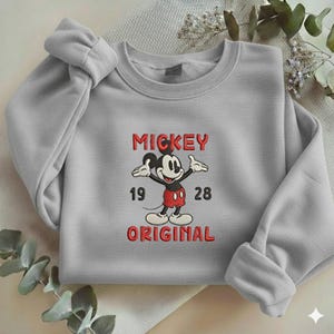 May include: A light gray crewneck sweatshirt featuring an embroidered graphic of Mickey Mouse in red shorts and yellow shoes. The text MICKEY is above the character, with 19 and 28 on either side, and the word ORIGINAL printed below in red.