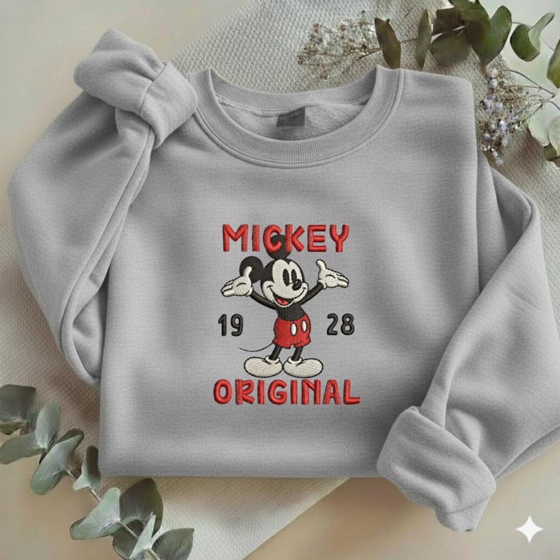 May include: A light gray crewneck sweatshirt featuring an embroidered graphic of Mickey Mouse in red shorts and yellow shoes. The text MICKEY is above the character, with 19 and 28 on either side, and the word ORIGINAL printed below in red.