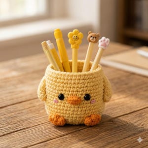 May include: A yellow crochet pen holder shaped like a chick, with black eyes, an orange beak and feet, and small wings. The holder contains yellow pens with decorative tops, including a flower, a bear, and a paw.