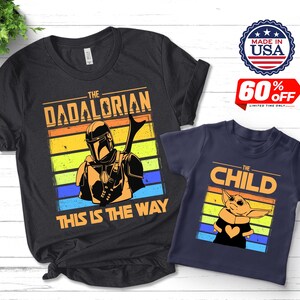 May include: Two t-shirts, one black and one navy blue, featuring graphics from a popular science fiction series. The black shirt says "The Dadalorian" with an illustration. The navy shirt says "The Child" with a character illustration. Both shirts have a retro sunset design.